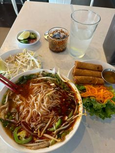 a bowl of soup with noodles, vegetables and dipping sauce on the side next to a glass of water