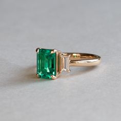 Lab grown emerald adorned with baguette diamonds Metal: 18K Yellow Gold Stone: Lab Grown Emerald Stone Weight: 1.4 Carat Stone Size: 8mm x 6 mm Accent Stone: Baguette Diamond Diamond Size: 4.5 x 2 mm (2) Diamond Color and Clarity: GHI, SI Total Diamond Weight: 0.25 TCW Gia Certified Baguette Cut Emerald Ring In 14k Gold, Gia Certified Baguette Cut Emerald Ring, Luxury Diamond Ring With Emerald In Baguette Cut, Luxury Emerald Diamond Ring With Baguette Cut, Luxury Three Stone Baguette Cut Rings, Elegant Emerald Ring With Baguette Diamonds, Luxury Moissanite Emerald Ring Baguette Cut, Baguette Cut Three Stone Emerald Diamond Ring, Luxury Moissanite Emerald Ring With Baguette Cut