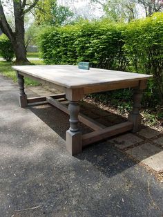 a wooden table sitting in the middle of a sidewalk next to a bush and shrubbery