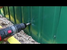 a man is using a drill to fix a green metal fence with rocks and gravel