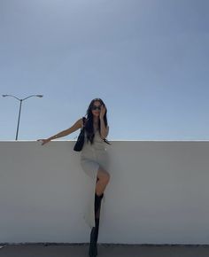 a woman leaning against a white wall with her legs crossed and one hand on her head