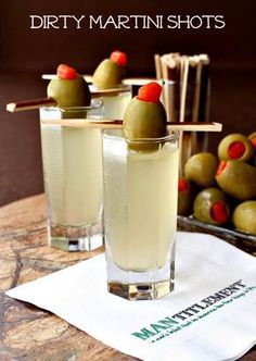 two glasses filled with liquid and olives on a table next to other food items