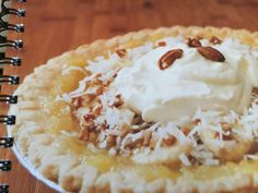 a pie topped with whipped cream and pecans on top of a wooden table next to a spiral notebook