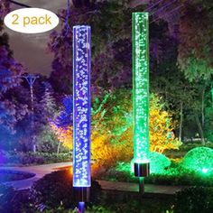 three lighted poles in the middle of a park at night with trees and bushes behind them