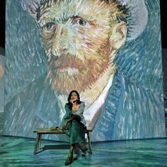 a woman sitting on top of a wooden bench in front of a large painting behind her