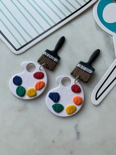 two paintbrush shaped earrings sitting on top of a table next to a pair of scissors