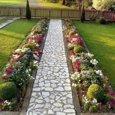 a stone path in the middle of a garden with flowers growing on each side and trees behind it