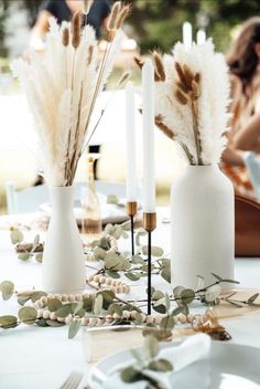 two white vases filled with feathers sit on a table next to candles and greenery