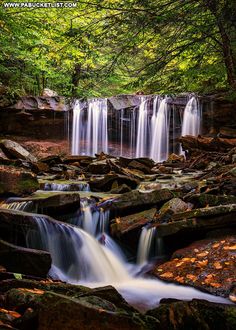 a small waterfall in the middle of a forest filled with rocks and leaves, surrounded by trees