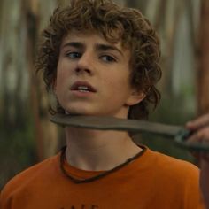 a young man with curly hair holding a knife in front of his face while wearing an orange shirt