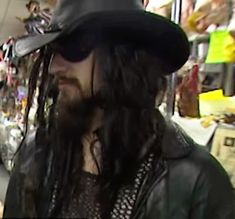 a man with long hair wearing a black hat and leather jacket in a market area