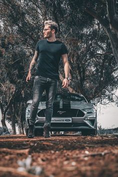 a man standing next to a car in front of some trees and dirt with one hand on his hip