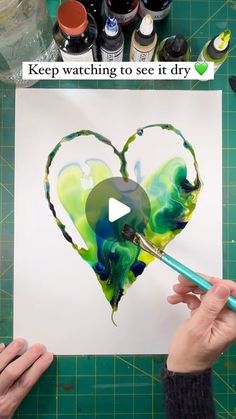 someone is painting a heart with green and blue colors on paper, while another person holds a paintbrush in their hand