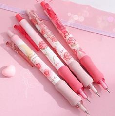 four pens are lined up on a pink surface next to an eraser and heart