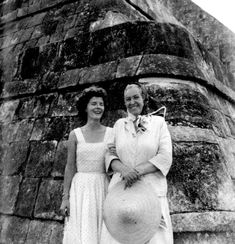 an old black and white photo of two women standing next to each other in front of a stone wall