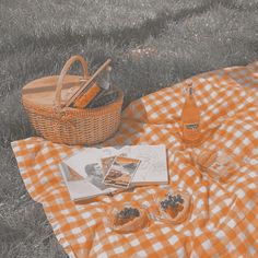 an orange and white checkered blanket on the grass next to a picnic basket with food