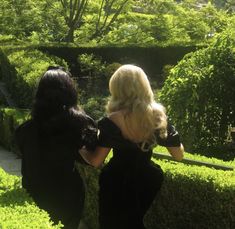 two women dressed in black are walking through the bushes and hedges together, looking at each other