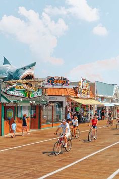 several people riding bikes on a boardwalk near shops and a large shark statue in the background