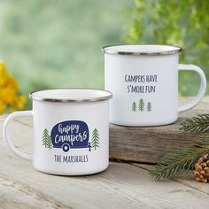 two white campers mugs sitting on top of a wooden table next to pine cones