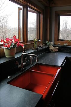 a kitchen with black counter tops and red sink in front of two large windows that look out onto the woods