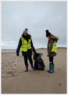 two women in yellow vests and boots on the beach with luggage bags, one holding crutches