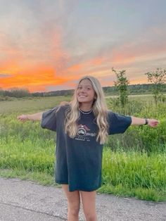 a woman standing on the side of a road with her arms outstretched in front of an orange sky