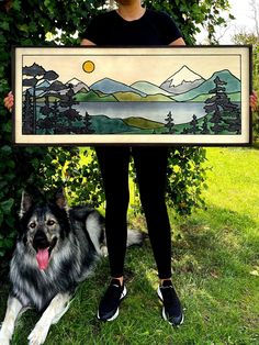 a woman holding up a painting with a dog laying next to her on the grass