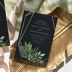 two black and gold wedding cards with greenery on them, next to a white satin ribbon