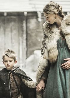 a woman holding the hand of a young boy who is wearing a fur coat and standing next to her