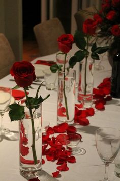 red roses are in vases lined up on a table with wine glasses and plates