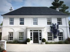 a large white house with two black doors and windows on the front of it, surrounded by greenery