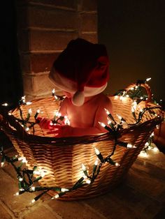 a baby is sitting in a basket with christmas lights on the floor and wearing a santa hat