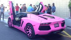 a pink sports car parked in front of a building with its doors open and people standing around