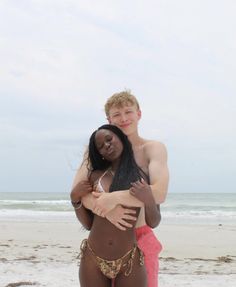 two people standing on the beach hugging each other and smiling at the camera, with one person wearing a bathing suit