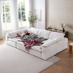 two people laying on top of a white couch in a living room with large windows