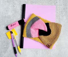 a crocheted hat and other crafting supplies on a pink surface with marker pens