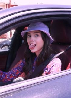 a woman sitting in the passenger seat of a car with her tongue out and wearing a hat