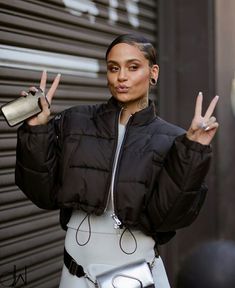 a woman in black jacket and white pants holding up two peace signs with one hand