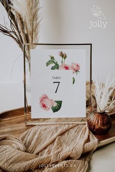 a table number card with pink flowers and greenery in front of it on a wooden tray