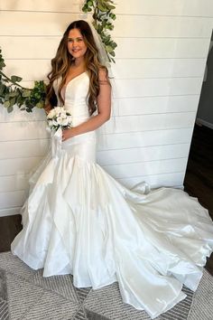 a woman standing in front of a white wall wearing a wedding dress and holding a bouquet