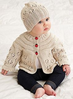 a baby sitting on a bed wearing a knitted jacket and hat with buttons in the front