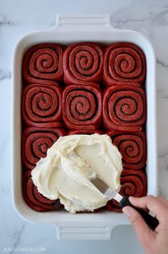 someone is spreading icing on top of red rolls in a baking dish with white frosting