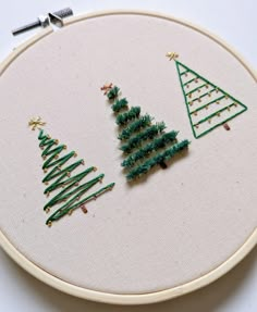 three small christmas trees are on a white embroidery hoop with green thread and gold stars