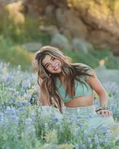 a beautiful young woman sitting on top of a field of blue flowers smiling at the camera