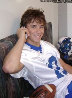 a young man sitting on top of a couch talking on a cell phone and holding a football