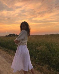 a woman standing on the side of a dirt road in front of a field at sunset