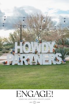 a large sign that says howdy partners in front of some trees and grass with lights above it