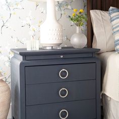 a night stand with two drawers and a lamp on it next to a flowered wallpaper