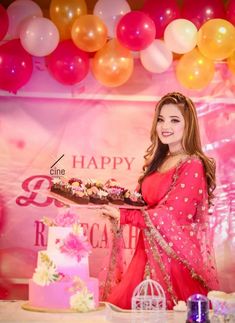 a woman in a red sari holding a cake with flowers on it and balloons behind her