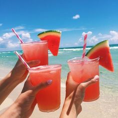 three people holding up glasses with watermelon juice on the beach in front of them